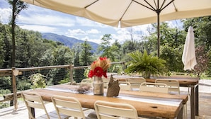 An outdoor dining area with a wooden table and white chairs, surrounded by a stone patio and a view of mountains. - Agriturismo Rossolampone (San Bernardino Verbano)
