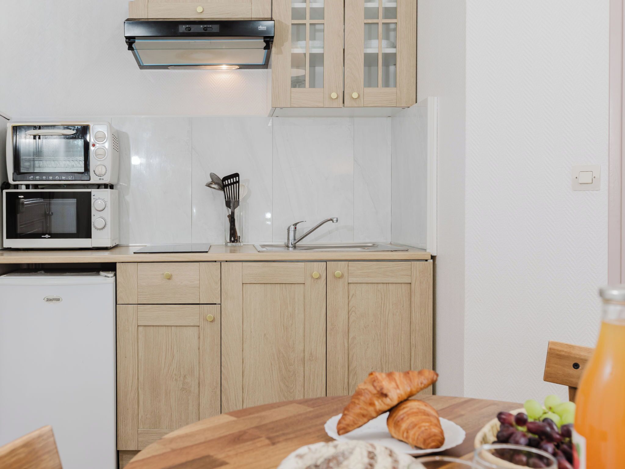 Une cuisine équipée d’un four à micro-ondes, d’un grille-pain et d’un lave-vaisselle intégrés, de placards en bois et d’une table à manger où sont servis des croissants et des raisins.