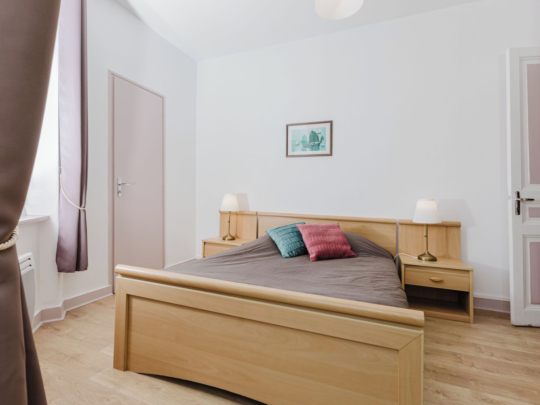 A bedroom with a wooden bed, bedside tables, a nightstand, a door, and a framed picture on the wall.