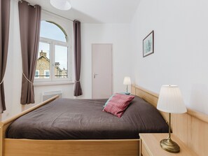 Une chambre à coucher avec un lit, une fenêtre avec des rideaux, une porte, une table de chevet avec une lampe et un tableau accroché au mur.
