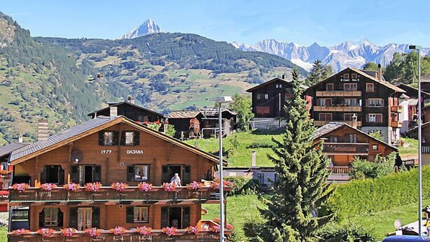 Un chalet traditionnel en bois, doté d’un balcon orné de fleurs, qui se détache sur un paysage de montagnes et d’un ciel dégagé.
