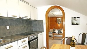 Une cuisine compacte dotée d’un four encastré, une table à manger avec une bouteille de vin, et un coin salon confortable avec un canapé et une lampe.