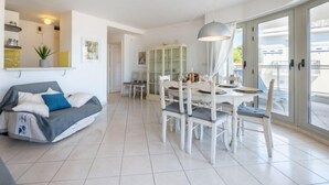 A modern living room with a sofa, dining table, and chairs. There is a cabinet with glass doors and a ceiling light fixture. - Les allées du Cap by Interhome (Antibes)