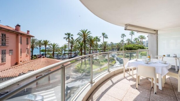 A balcony with a glass railing overlooking a street with parked cars and palm trees. - Les allées du Cap by Interhome (Antibes)