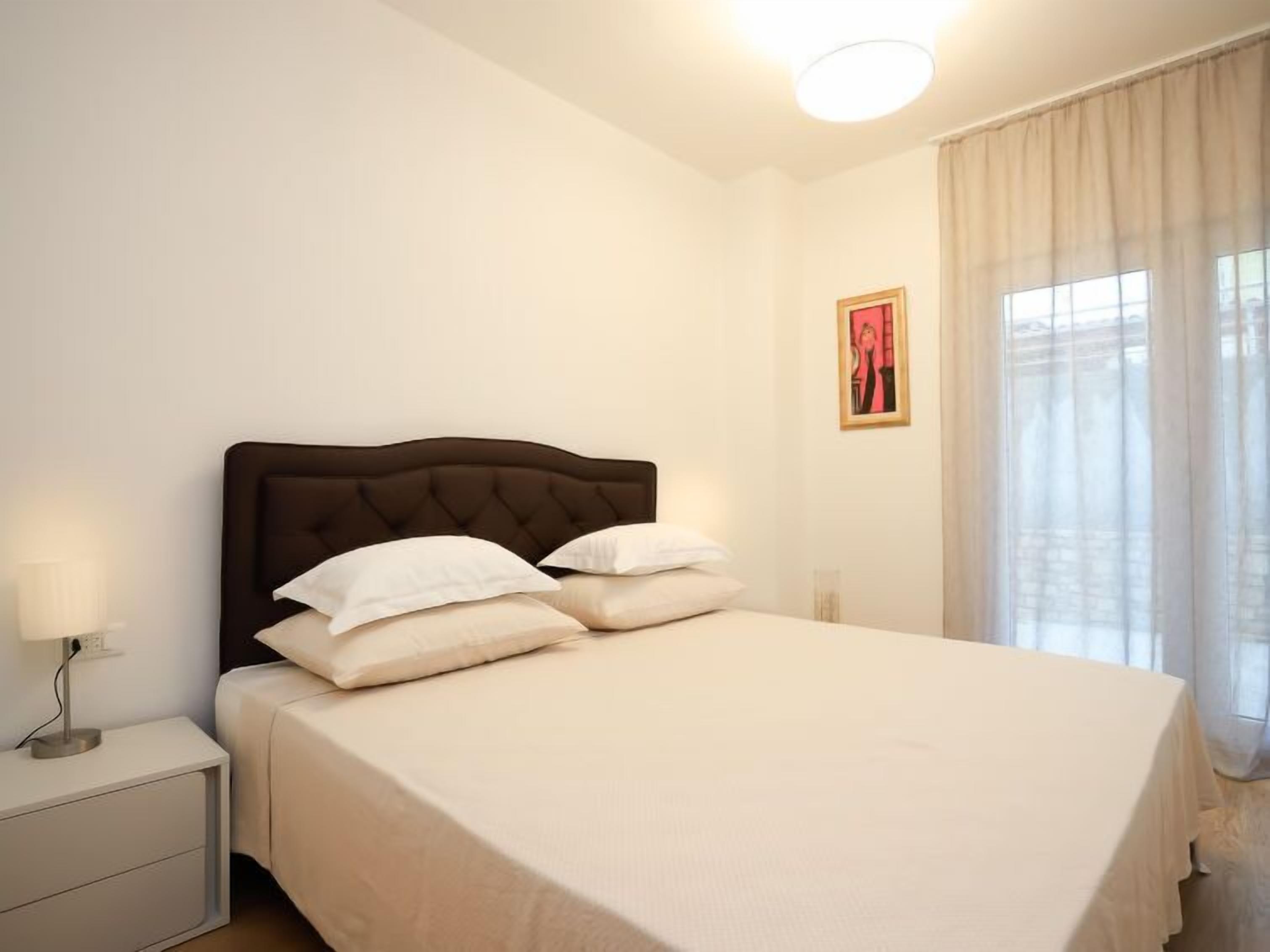Ein ordentlich bezogenes Bett mit dunklem Kopfteil in einem Zimmer mit weißen Wänden, einem Nachttisch und einem Fenster mit durchsichtigen Vorhängen.