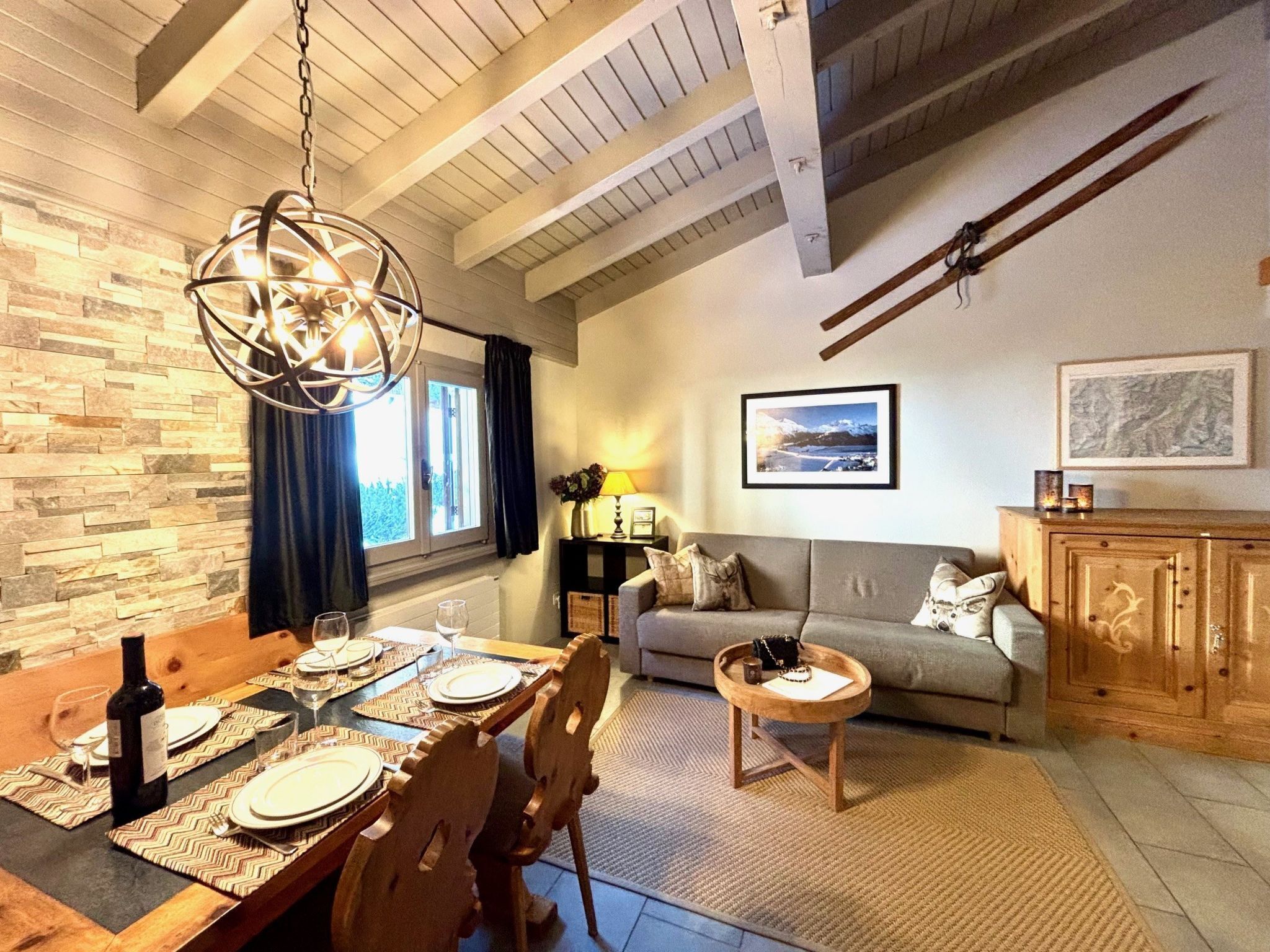 A cozy living room with a dining area, wooden furniture, a fireplace, and a large window with curtains.
