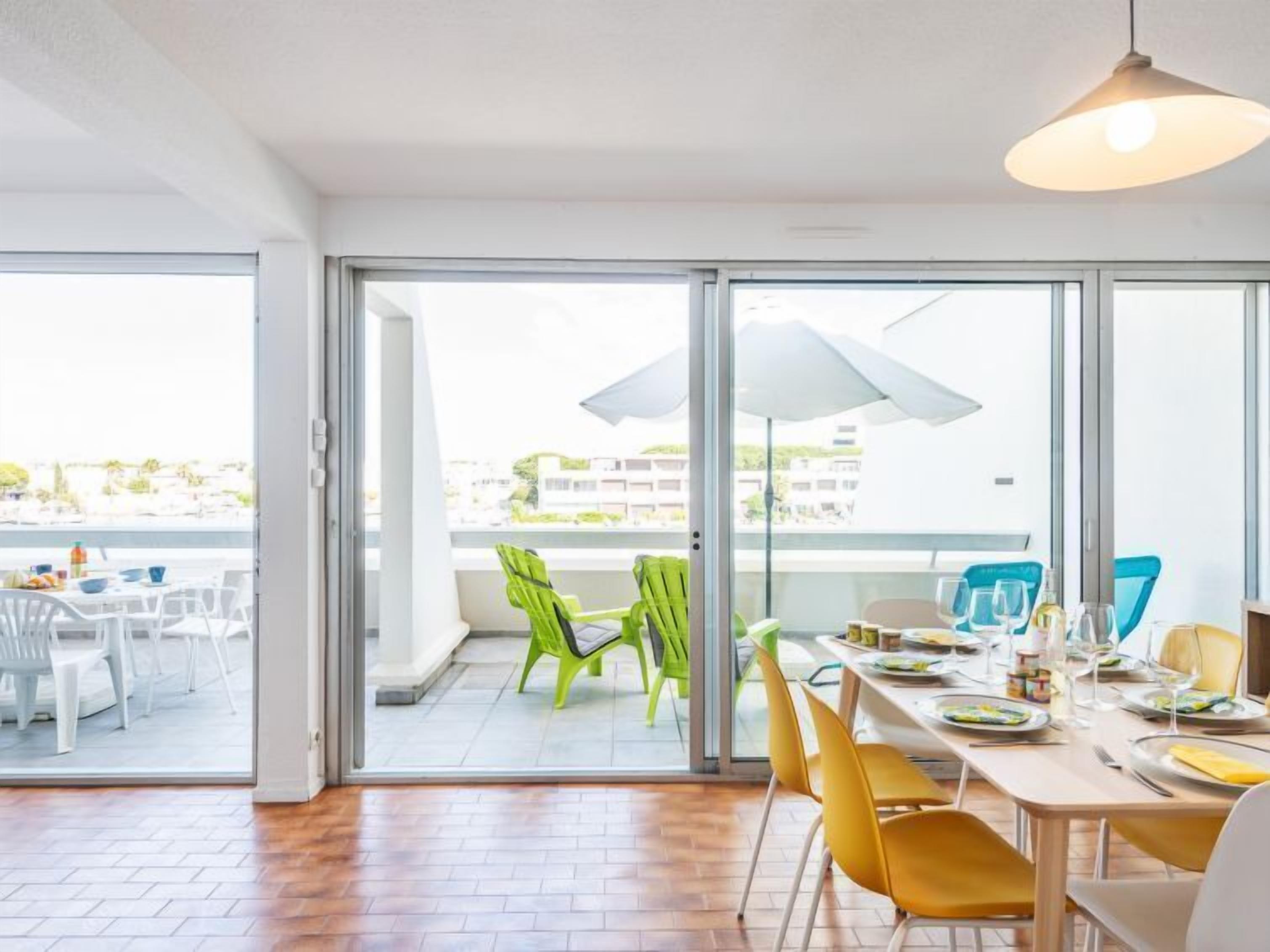Un comedor luminoso con una mesa grande dispuesta para cuatro personas, una puerta corredera de vidrio que da a un área de estar exterior, y una vista de la ciudad al fondo.