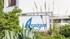 Une enseigne avec le mot « Arpèges » écrit en lettres bleues, entourée de verdure et d’un bâtiment blanc doté d’une fenêtre et de volets.