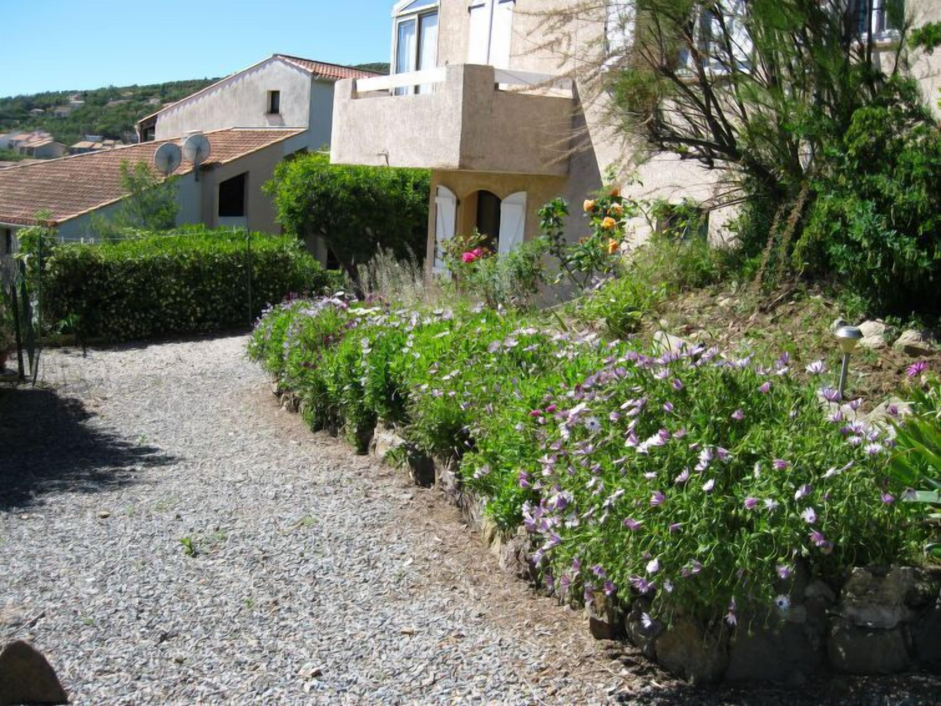 A gravel path leading to a house with a sloped roof, surrounded by lush greenery and flowering plants.