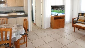 Een woonkamer met een televisie, een eettafel met stoelen, een bank en een keuken met kastjes en een spoelbak.