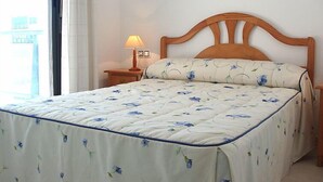 Een net opgemaakt bed met een bloemenprint beddeken, een houten hoofdbord en een bedlamp op een nachtkastje.