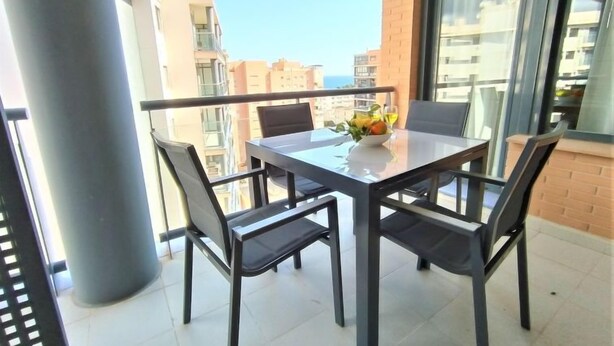 Een balkon met een tafel en stoelen, een vaas met bloemen en uitzicht op gebouwen en de zee.