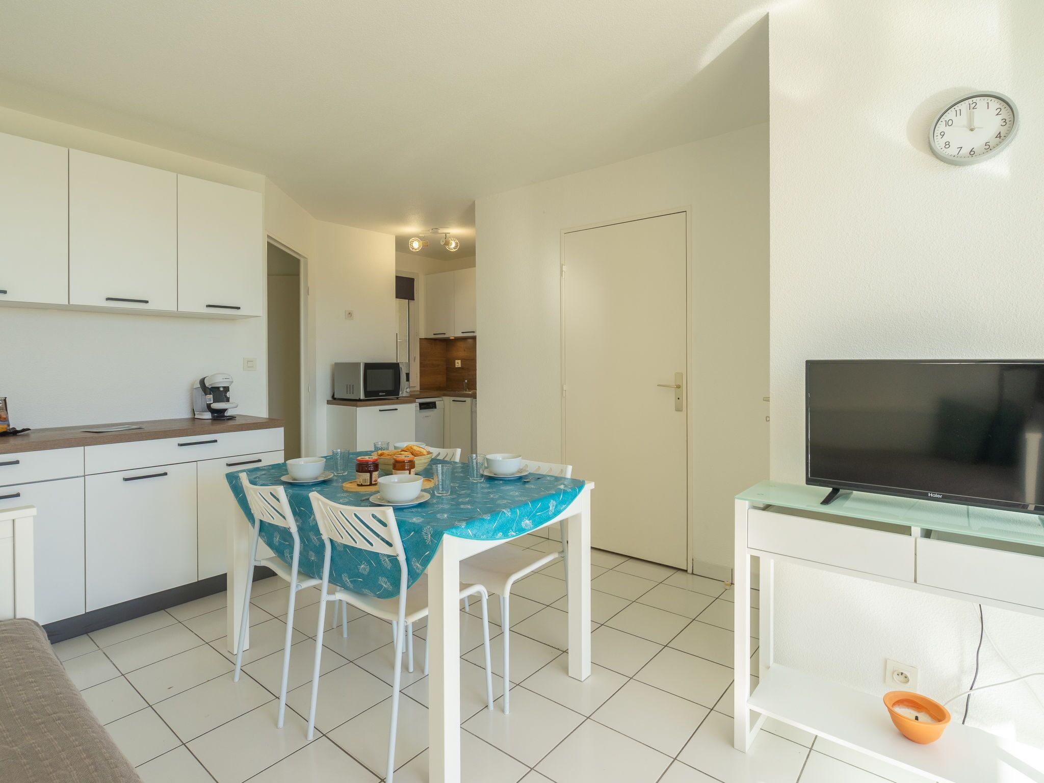 A compact kitchen with white cabinets, a dining table with chairs, a microwave, and a television on a stand.