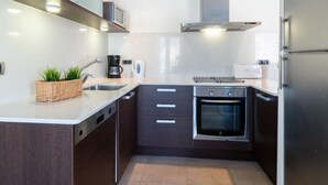 Een moderne keuken met een roestvrijstalen koelkast, een ingebouwde oven en een magnetron. Er is een spoelbak met een kraan, een koffiezetapparaat en een mand met groene planten. De kastjes zijn van donker hout, en het aanrecht is wit.