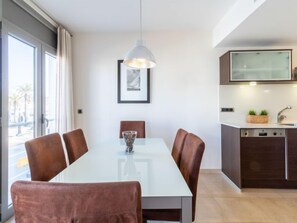 A modern dining area with a white table, brown chairs, and a pendant light. There is a kitchen with dark cabinets and a dishwasher. A framed picture hangs on the wall.
