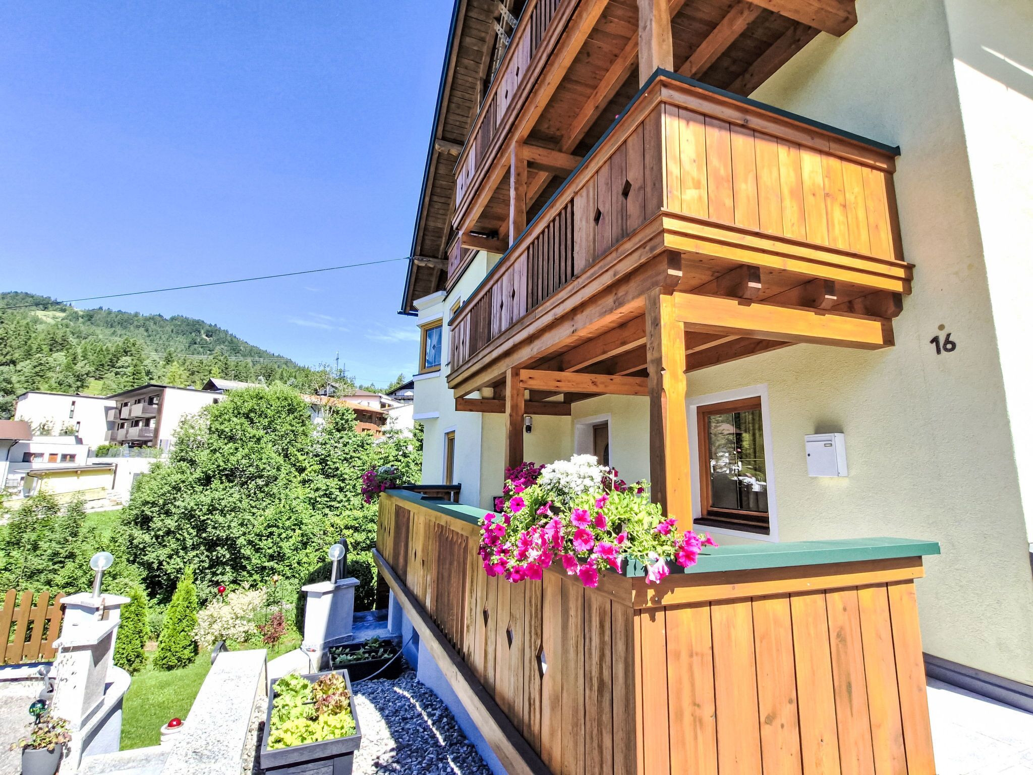 Un balcón de madera con jardineras de flores, un pequeño jardín y vistas a una colina.