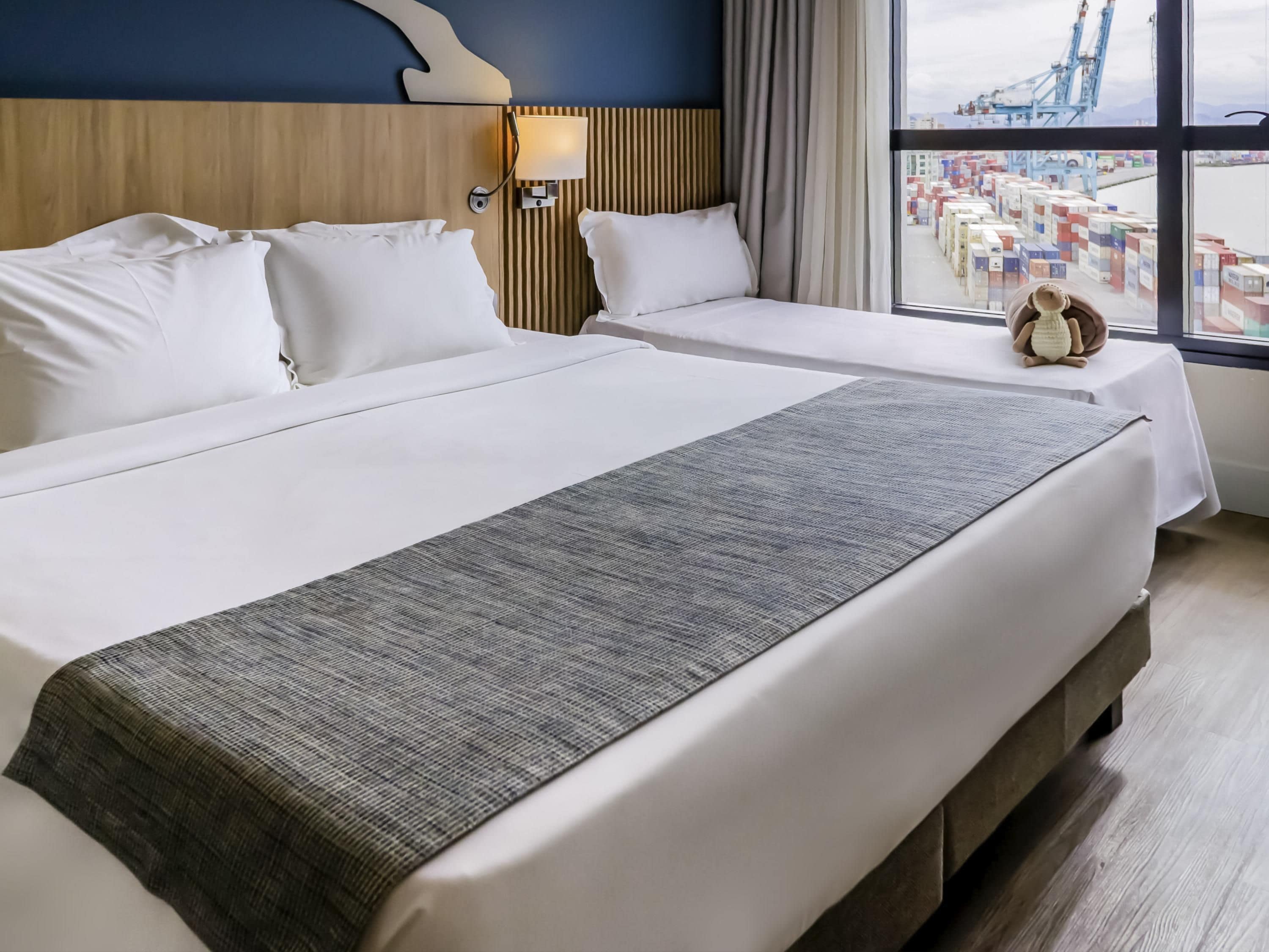 Et hotelværelse med en stor seng, en senelampe og udsigt til en havn med containere.