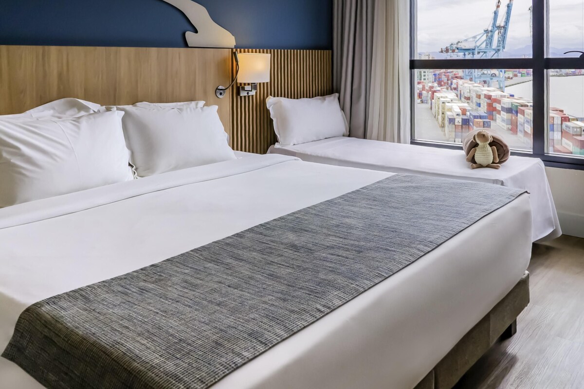 Une chambre d’hôtel avec un grand lit, une lampe de chevet et une vue sur un port où se trouvent des conteneurs.