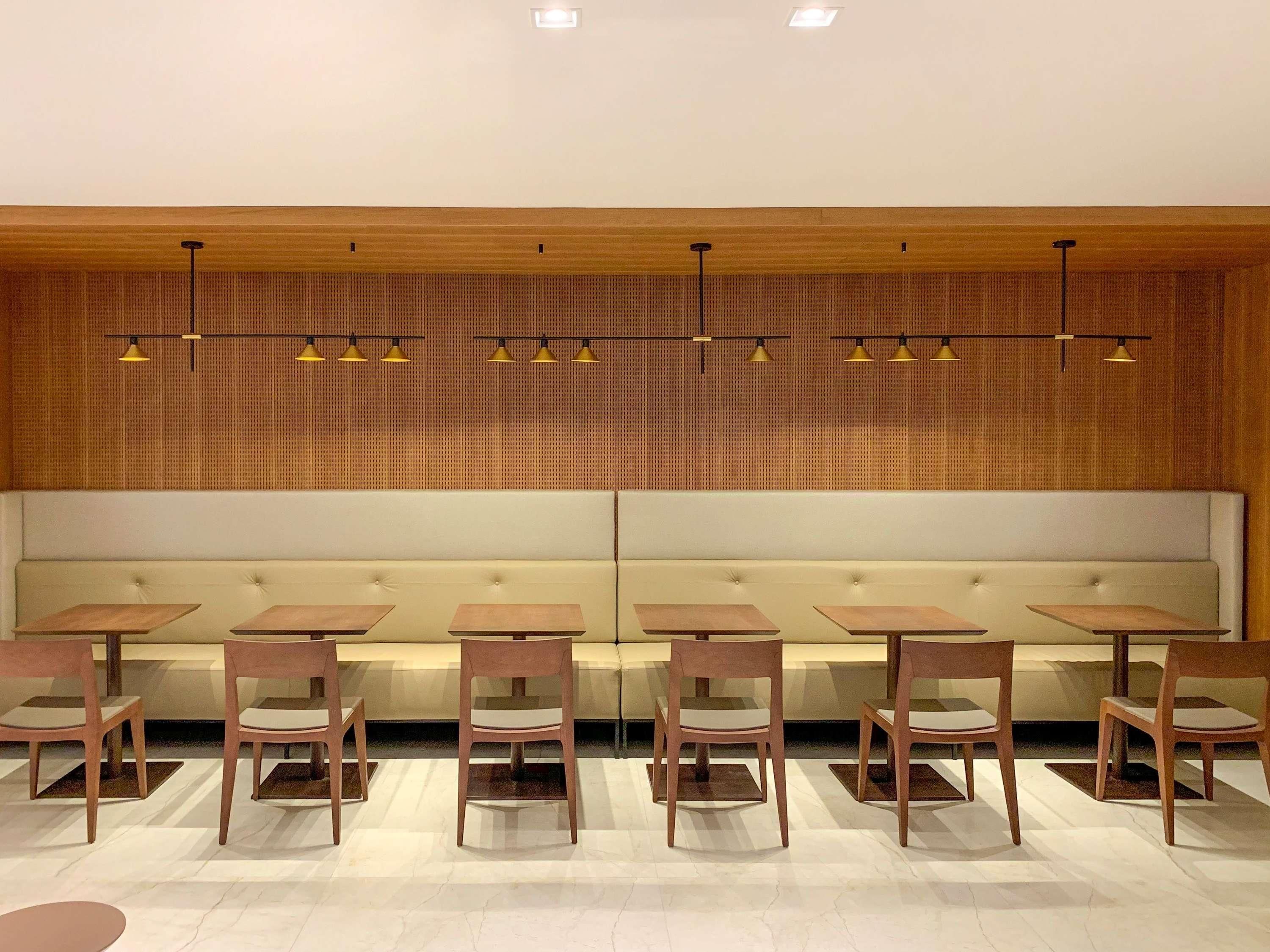 A modern interior with a row of wooden tables and chairs, a padded bench, and a patterned wall with recessed lighting.