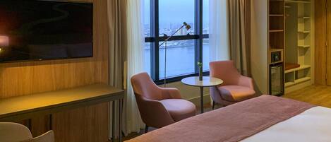 Habitación de hotel con cama, televisor montado en la pared, dos sillones, una mesita y vistas a un paisaje urbano a través de un amplio ventanal.