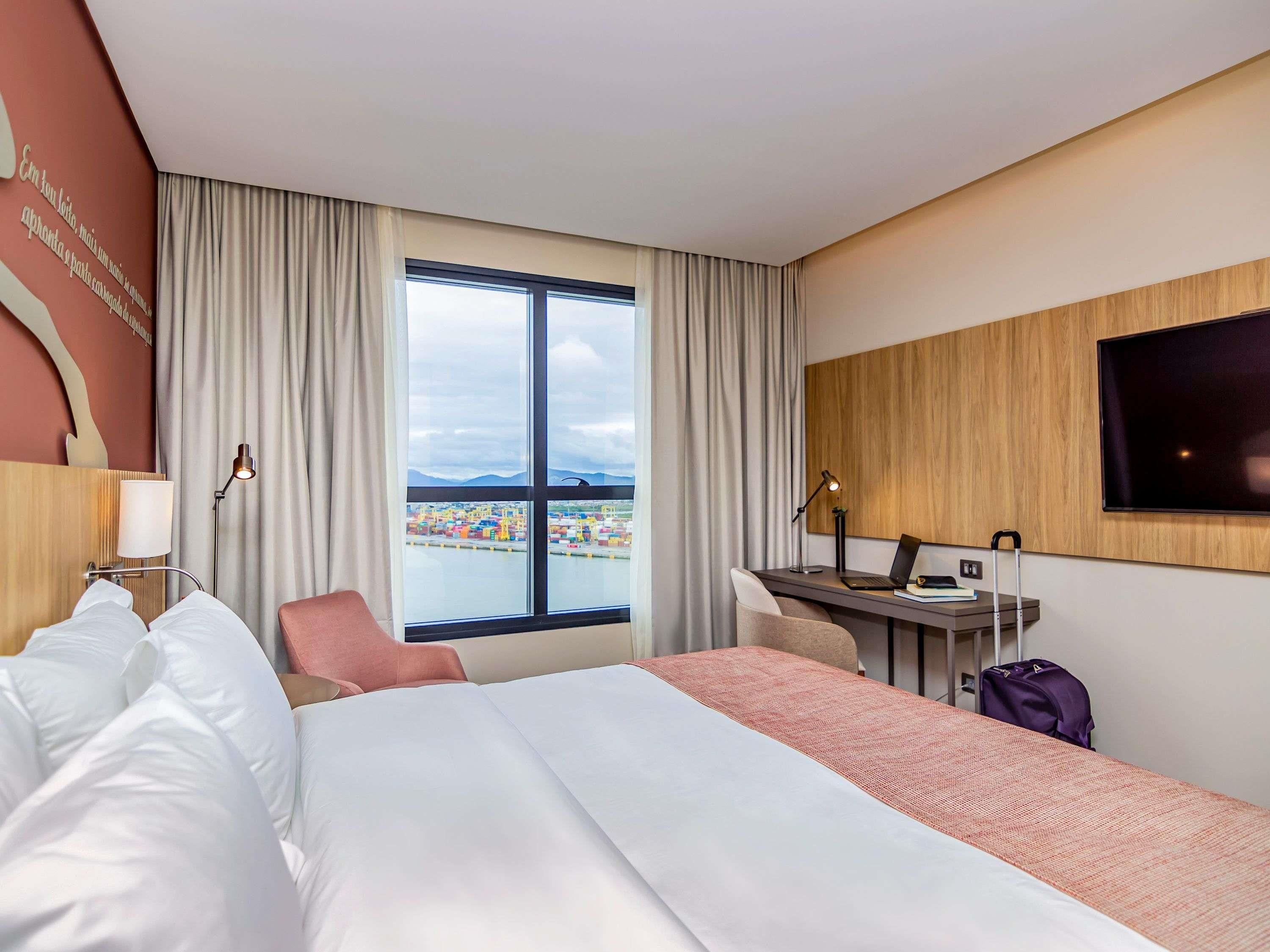 A hotel room with a bed, desk, chair, TV, and a large window with a view of a port.