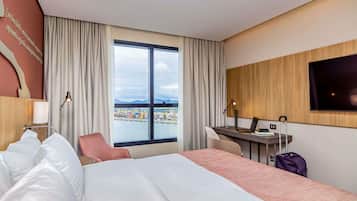 A hotel room with a bed, desk, chair, TV, and a large window with a view of a port.
