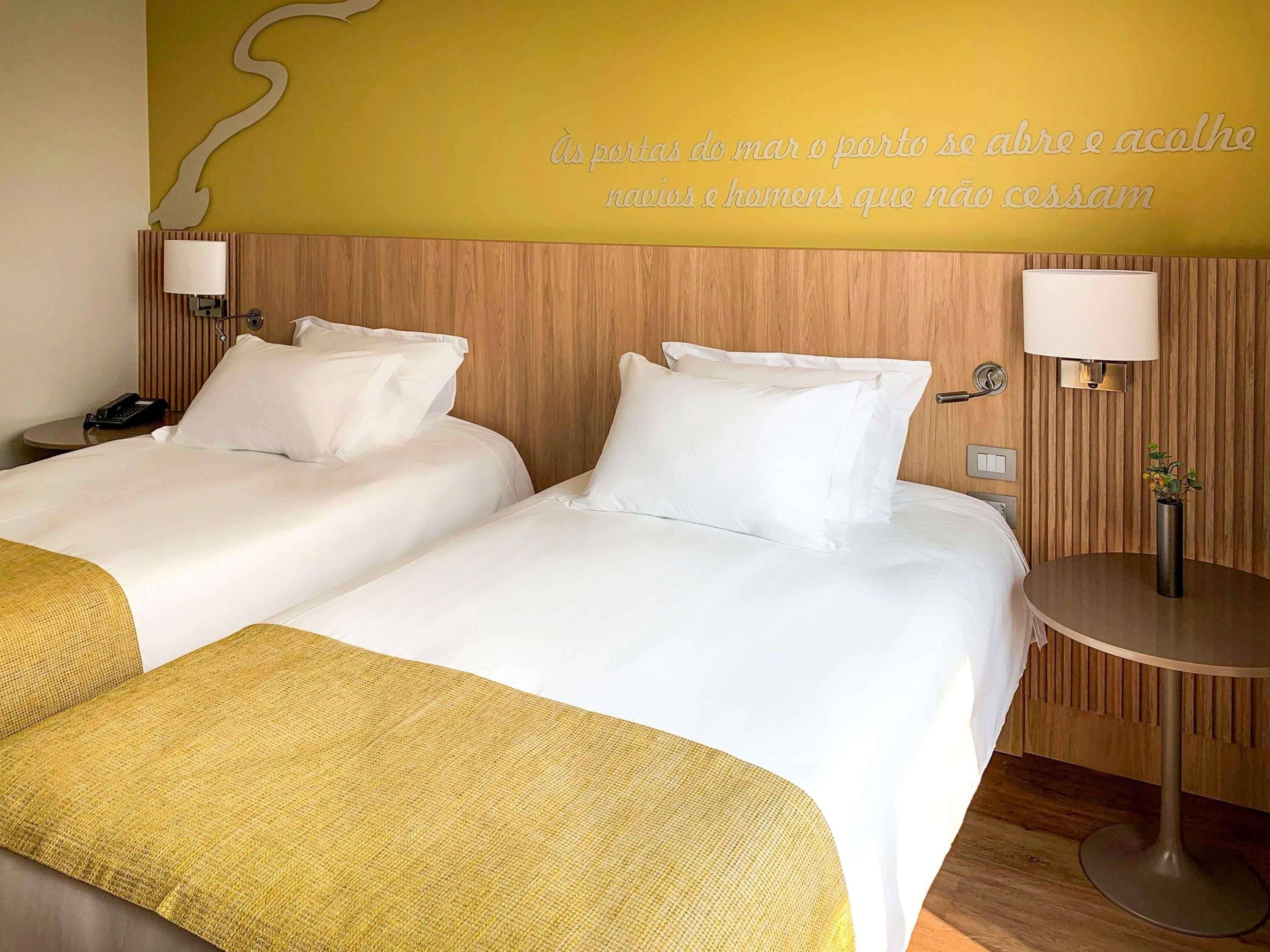 Une chambre d’hôtel avec deux lits, une tête de lit en bois, des lampes de chevet, une petite table avec un vase et un mur orné d’une citation décorative.