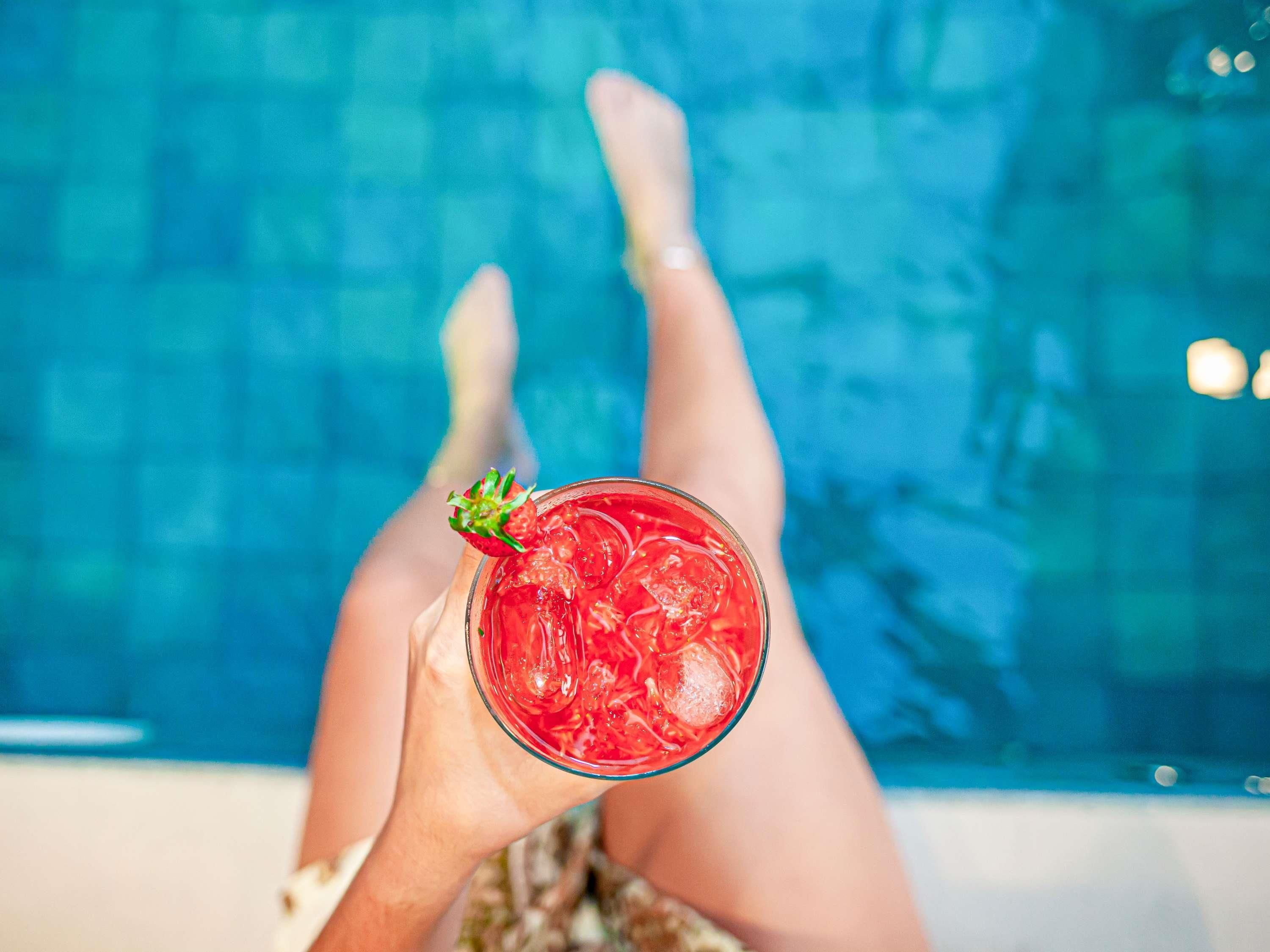 A person relaxing by a pool with a glass of red beverage and ice cubes.