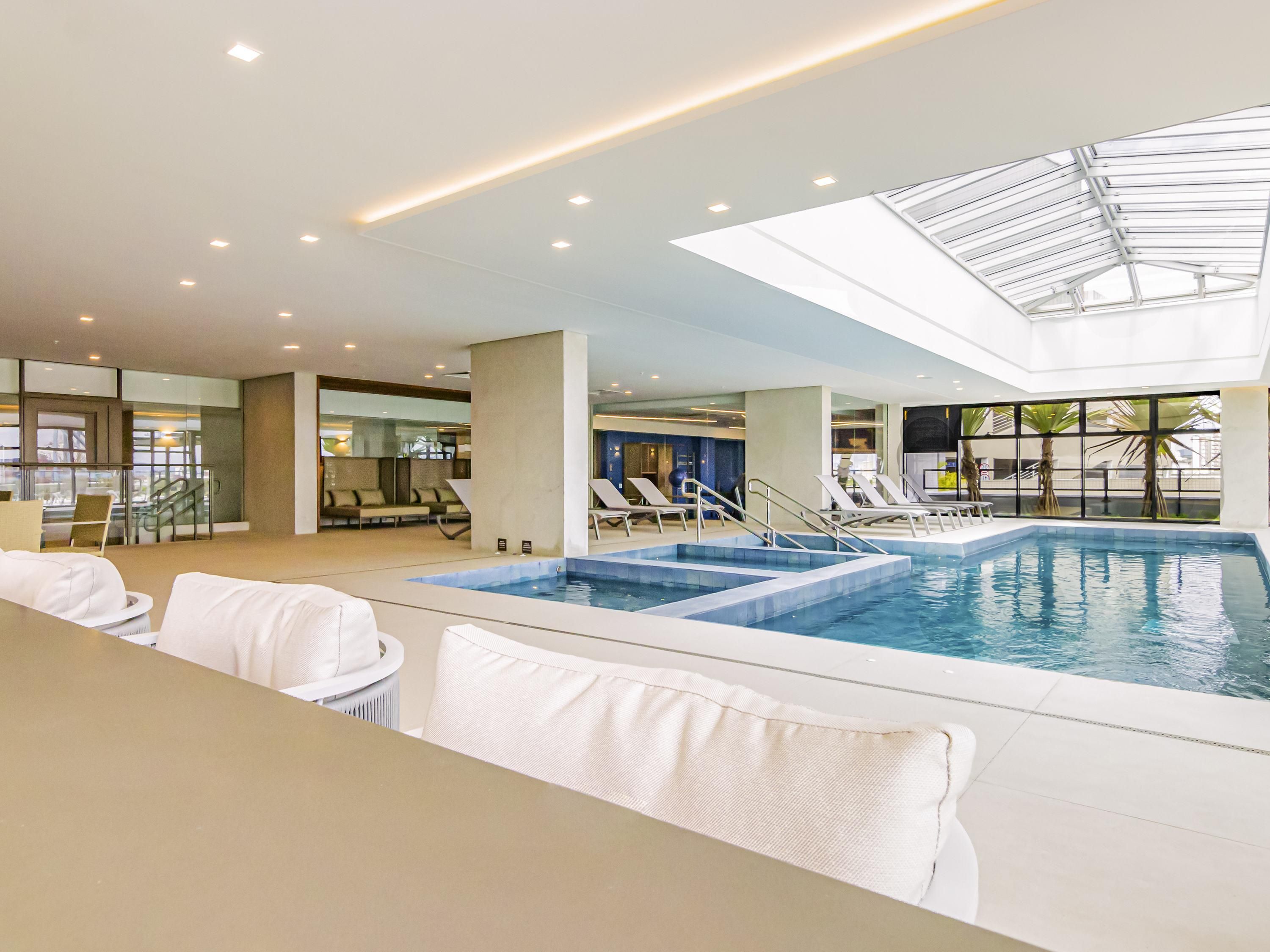 Una piscina interior con sillas de descanso, techo de vidrio y vistas al exterior.