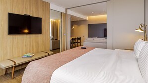 Una habitación de hotel moderna con una cama grande, un televisor montado en la pared, una zona de estar con un banco y un comedor separado.