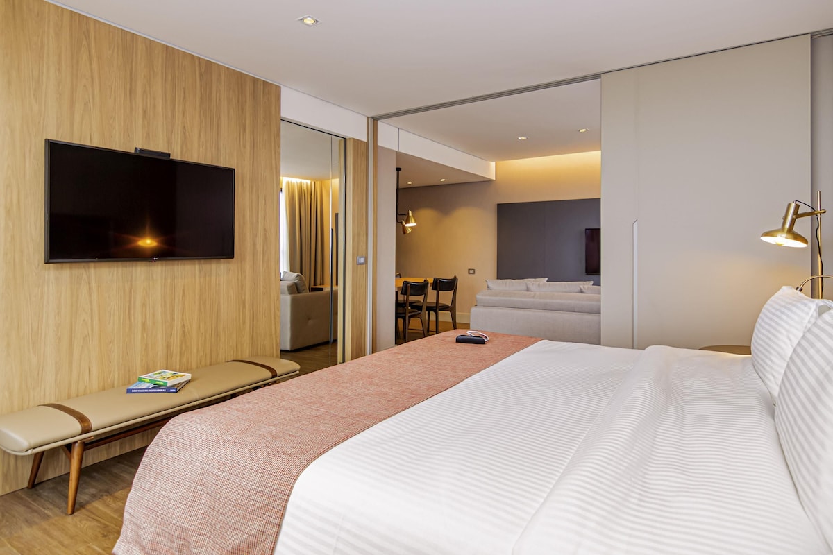 Una habitación de hotel moderna con una cama grande, un televisor montado en la pared, una zona de estar con un banco y un comedor separado.
