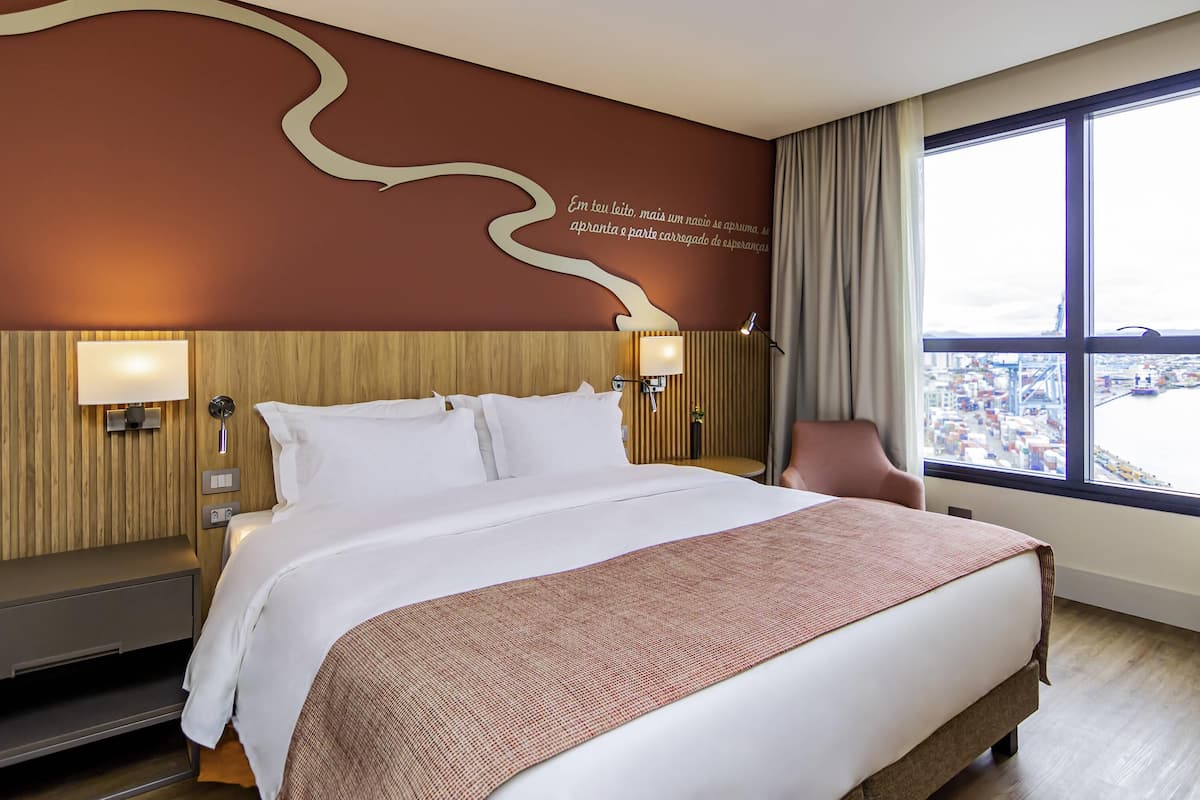 A hotel room with a large bed, a desk, a chair, a window with a city view, and a wall with a decorative design.