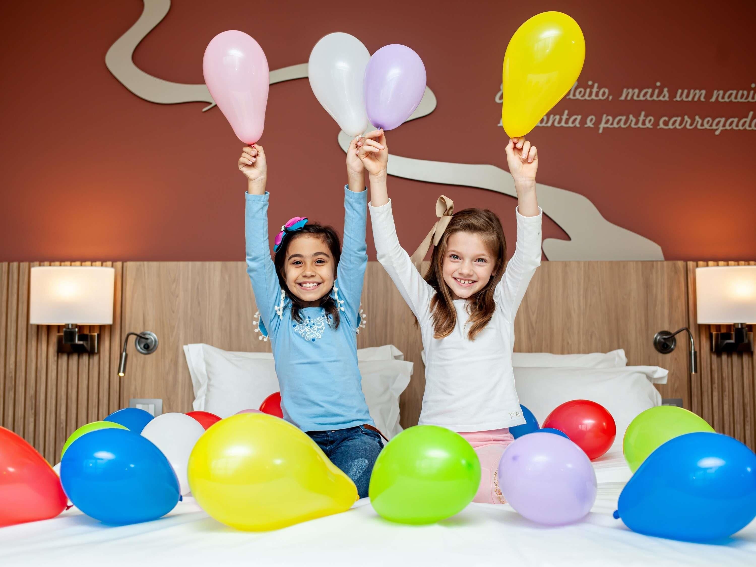 Twee personen zitten op een bed, omringd door kleurrijke ballonnen, in een kamer met houten lambrisering en wandlampen.
