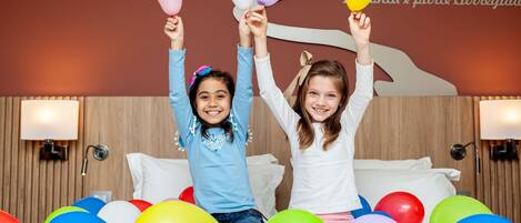 Twee personen zitten op een bed, omringd door kleurrijke ballonnen, in een kamer met houten lambrisering en wandlampen.