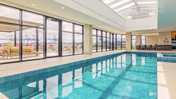 Ein Innenpool mit großen Glaswänden, Liegestühlen und Blick auf den Hafen.