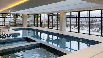 Piscina interior moderna com paredes de vidro transparente, área de hidromassagem separada e espreguiçadeiras.