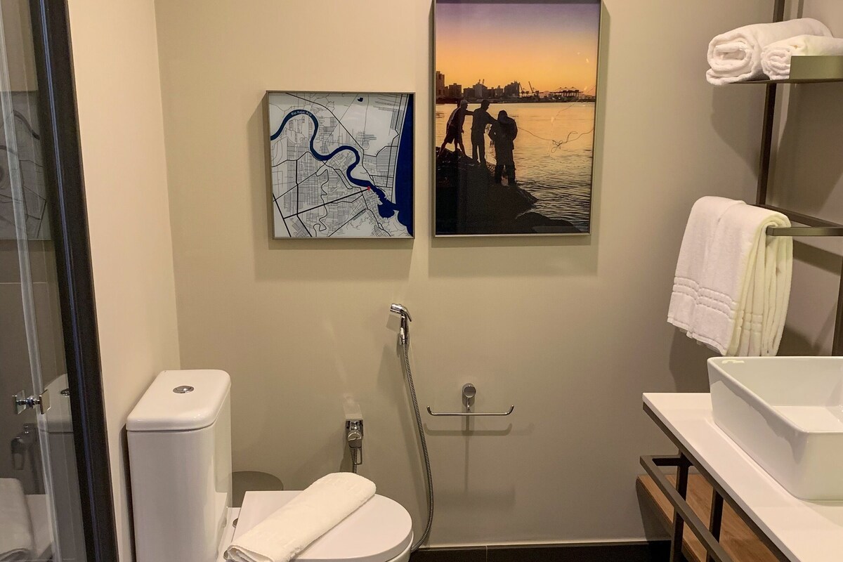 Un baño moderno con inodoro, bidé y ducha. Hay dos cuadros en la pared, uno de un mapa y otro de un paisaje al atardecer.
