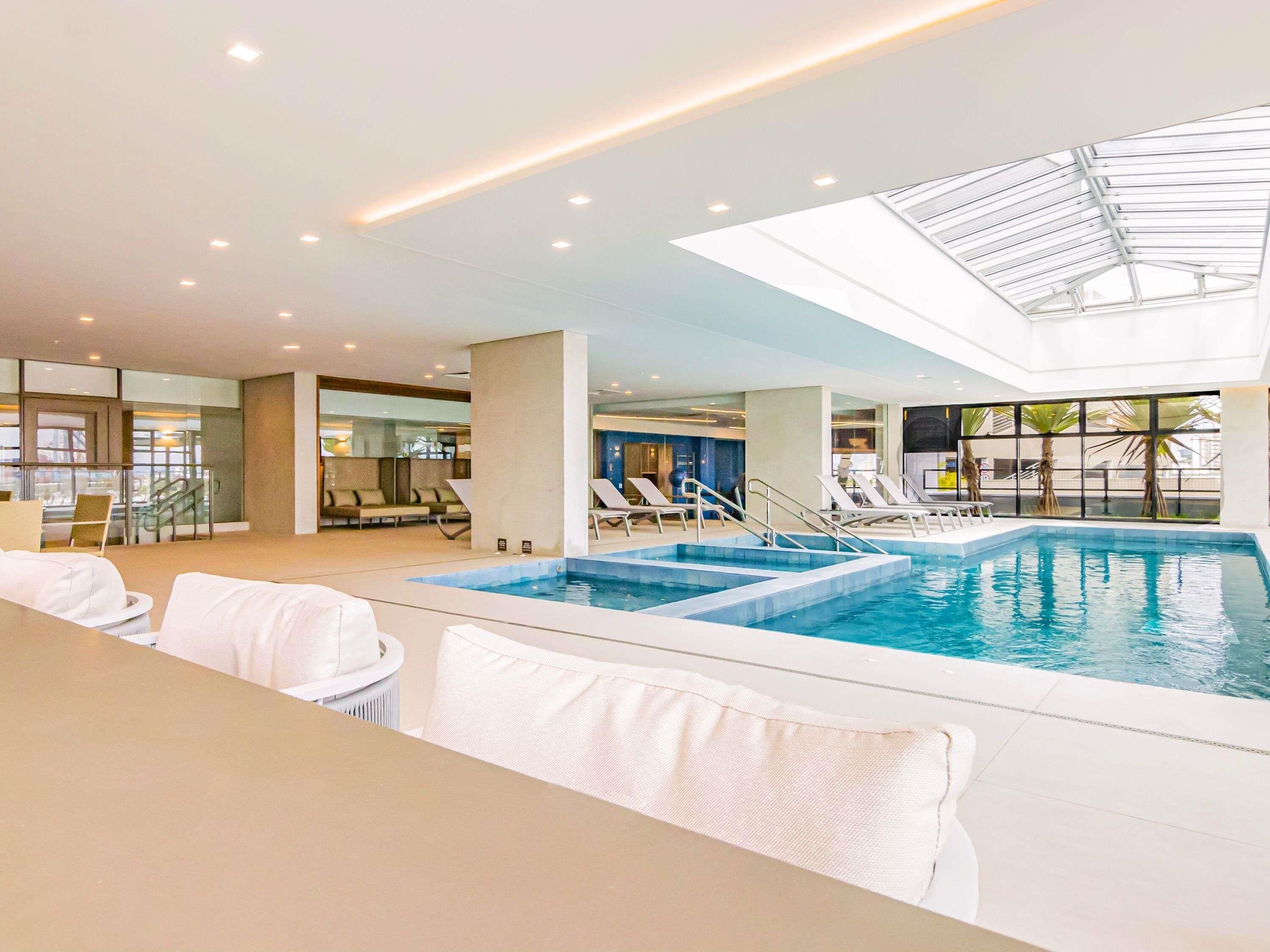 A swimming pool with sun loungers, a glass ceiling, and a view of the outside.