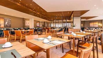 A spacious dining area with multiple tables and chairs, a high ceiling with wooden beams, and large windows.