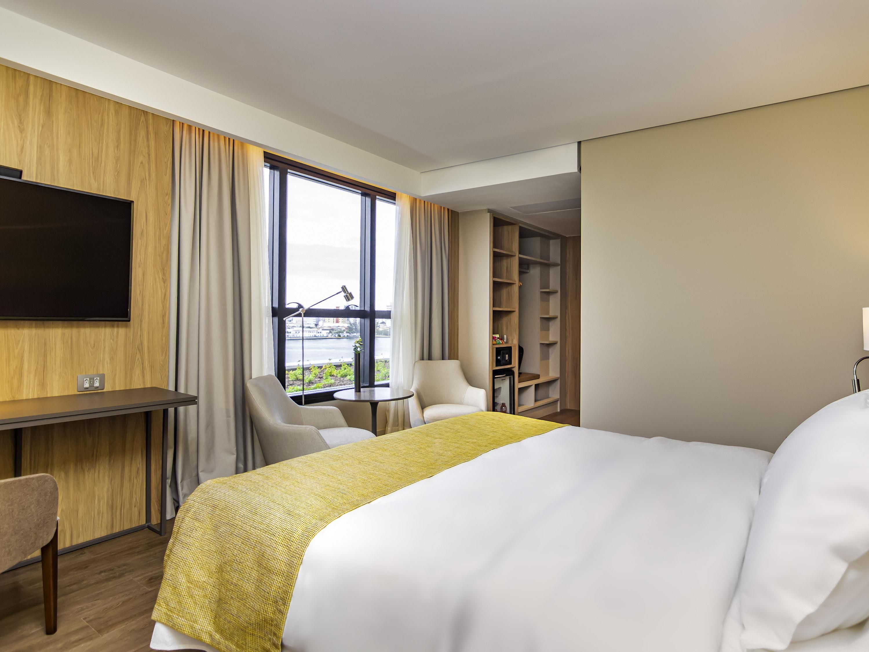 Une chambre d’hôtel comprenant un lit, un bureau, une chaise, une petite table, un téléviseur et une vue sur une étendue d’eau grâce à de grandes fenêtres.