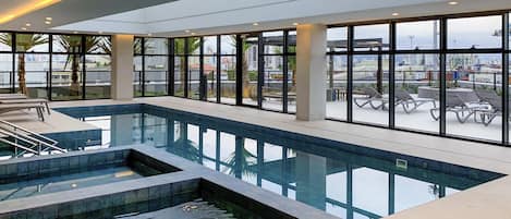 A modern indoor swimming pool with clear glass walls, a separate hot tub area, and lounge chairs.
