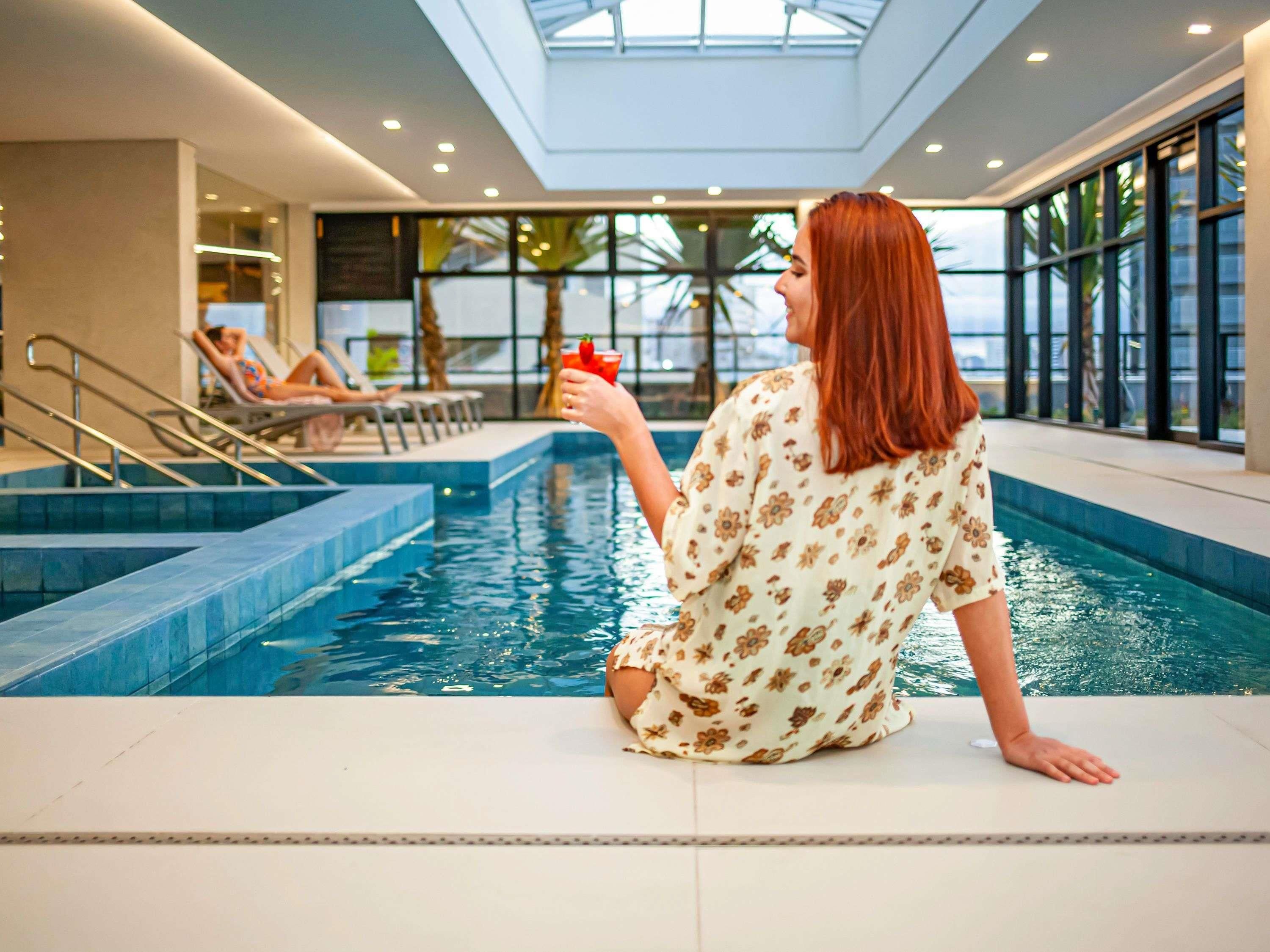 Piscina interna com teto de vidro, uma pessoa sentada na borda e outra relaxando em uma espreguiçadeira.