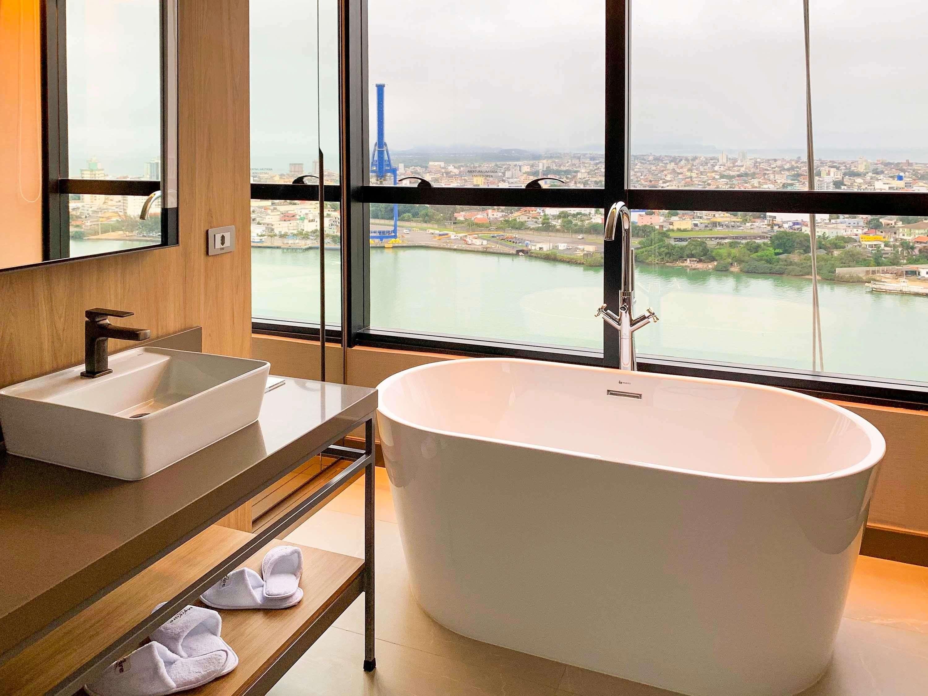 A modern bathroom with a freestanding bathtub, a sink, and a large window offering a view of a cityscape and a river.