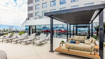 En tagterrasse med liggestole og en sofa, store vinduer med udsigt over en havn og kraner, samt en moderne bygningsfacade.