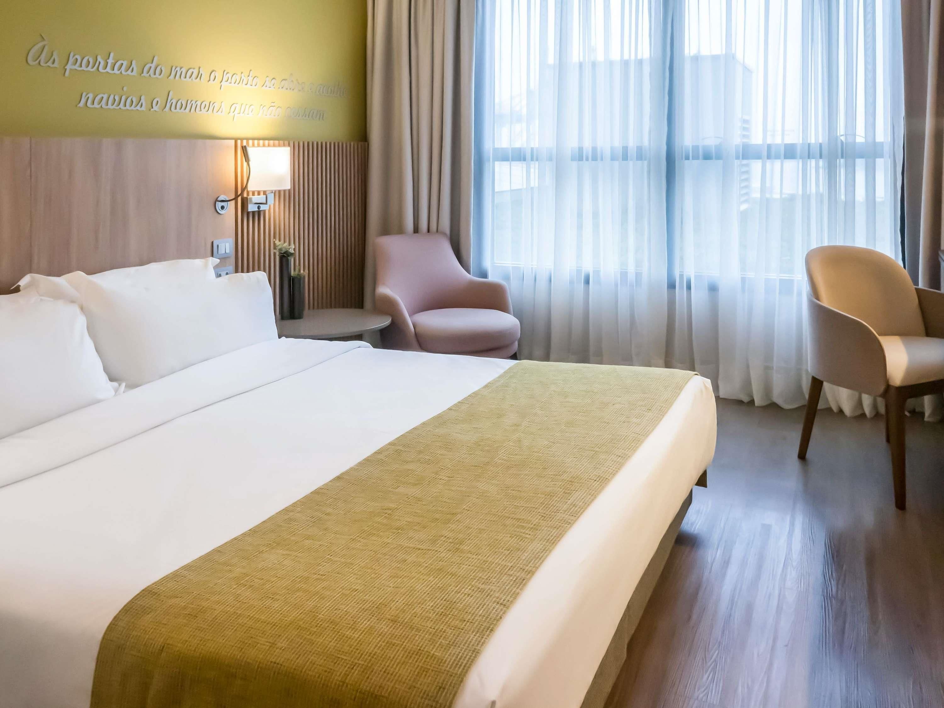 Ein Hotelzimmer mit einem großen Bett, einem Nachttisch mit Lampe, einem Sessel und Blick auf die Stadt durch transparente Vorhänge.