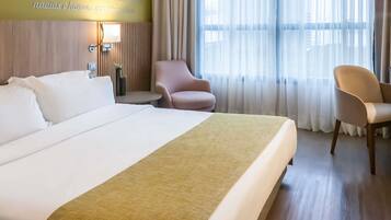 Ein Hotelzimmer mit einem großen Bett, einem Nachttisch mit Lampe, einem Sessel und Blick auf die Stadt durch transparente Vorhänge.