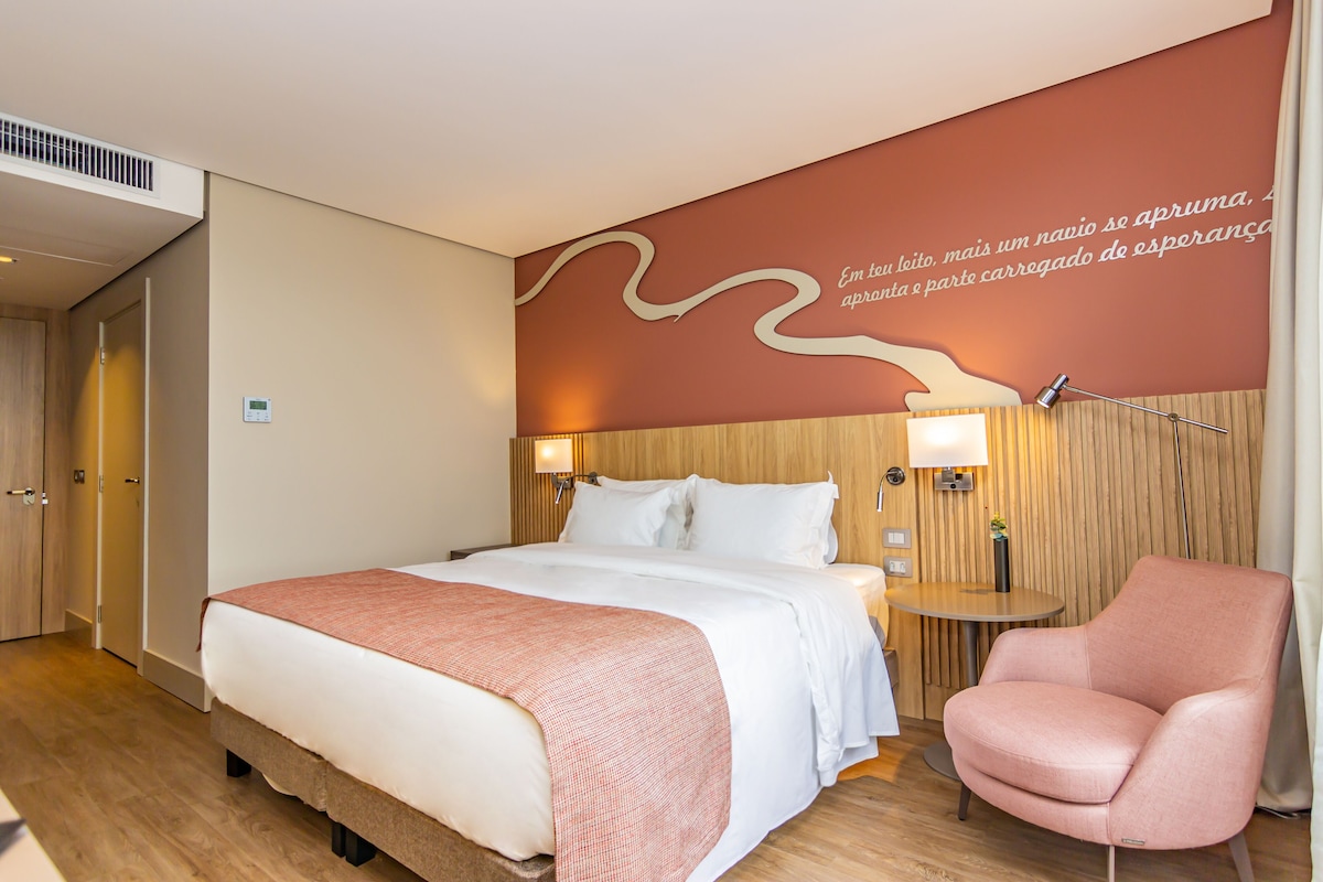 Habitación de hotel con cama, una silla rosa, una mesita con lámpara y una pared con una cita.