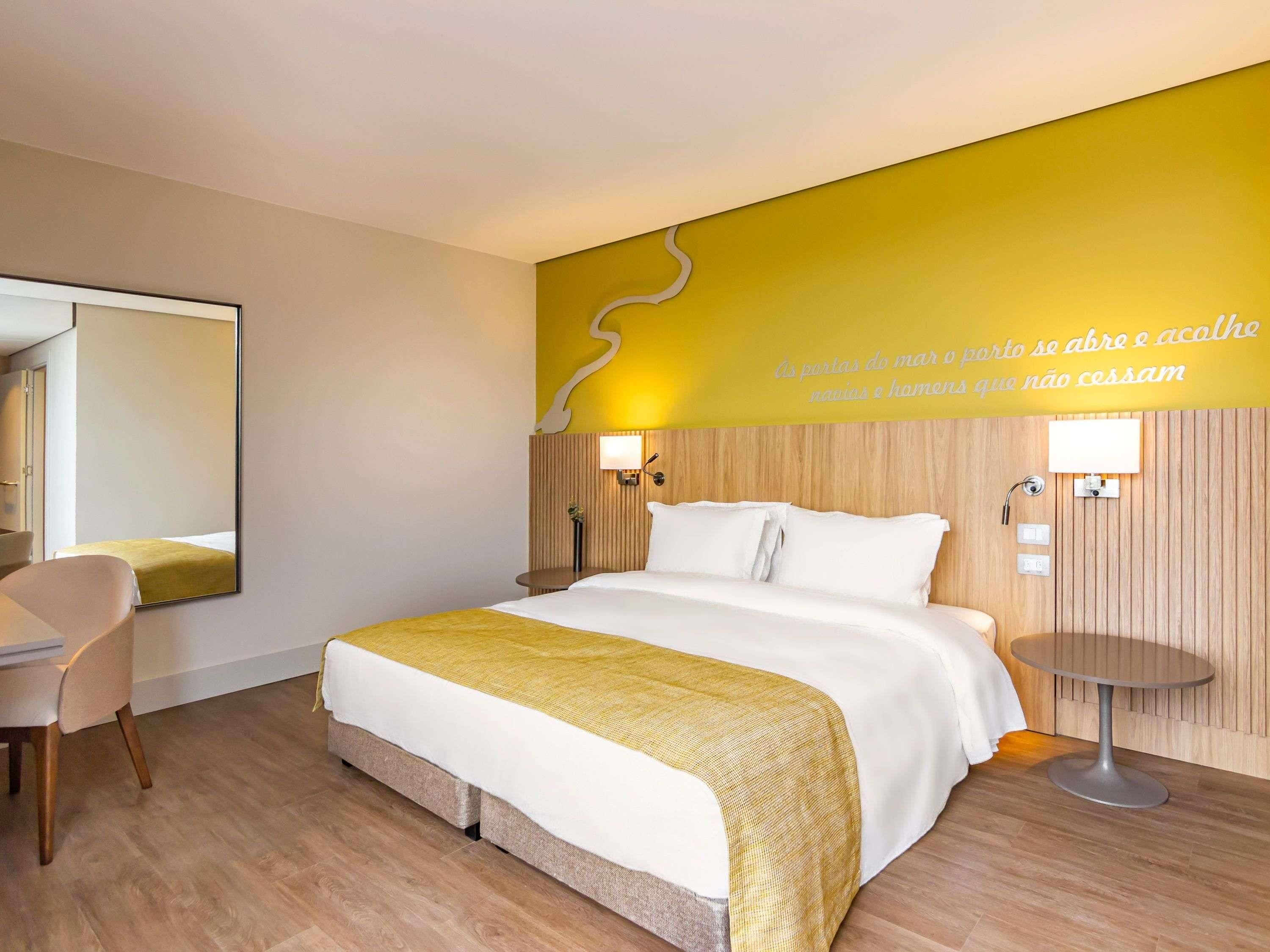 A hotel room with a bed, bedside table, chair, and a wall with a decorative element and text.