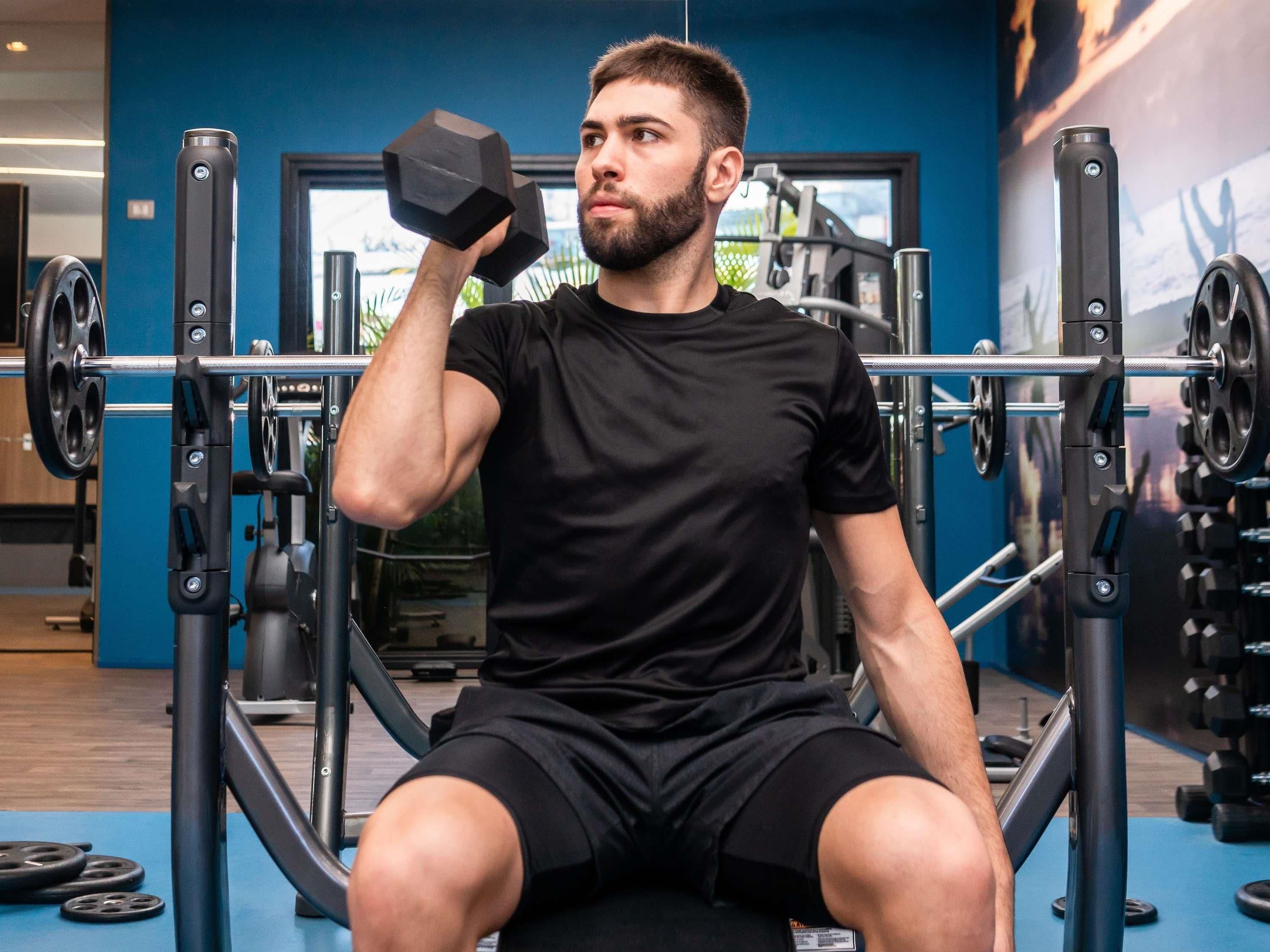 Una persona sentada en una máquina de gimnasio, con una barra y pesas de fondo.