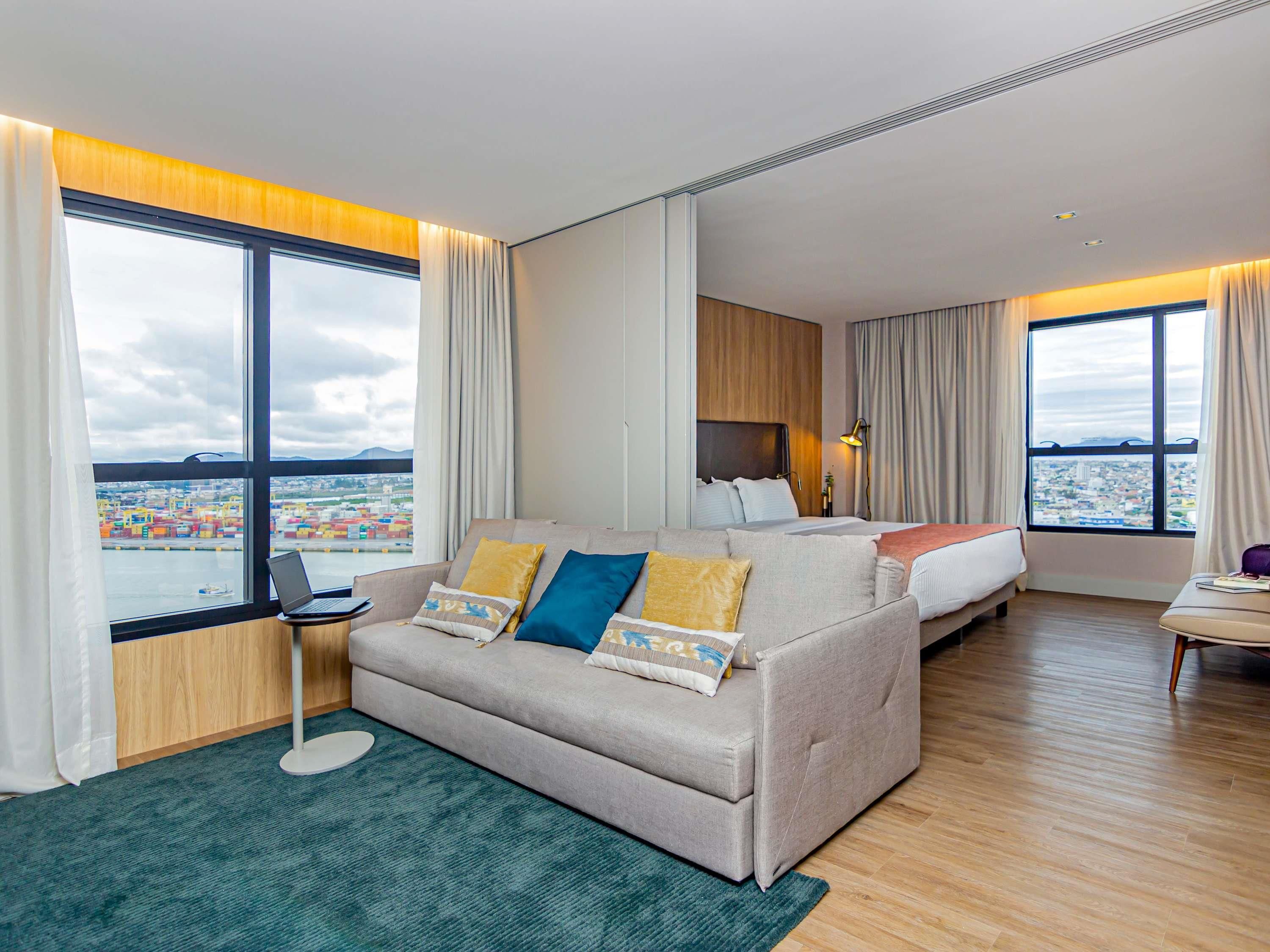 A modern hotel room with a large bed, a sofa, a desk, and a view of a cityscape.